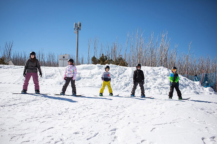 five kids standing on top of the hill at lake mountain strapped into a snowboard